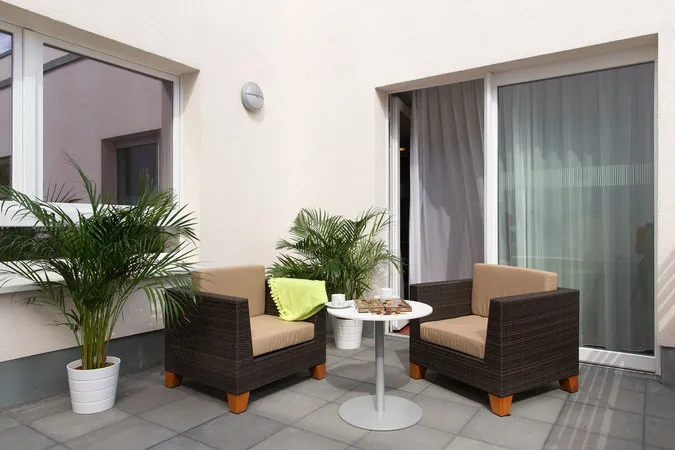 Zwei Korbsessel und ein Tisch auf modernem Balkon, mit Topfpflanzen und Vorhängen.